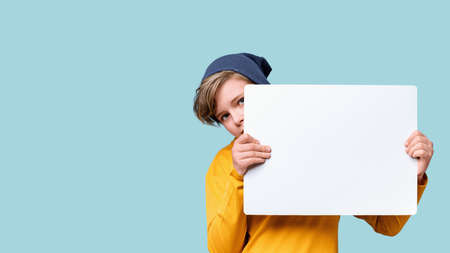 Young teen caucasian boy peeking out from behind white notice board with copy space. Blue background...