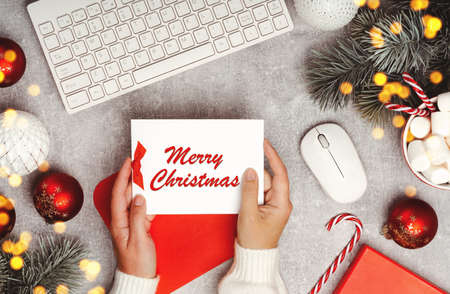 Hands holding a greeting card with Merry Christmas text. Red and white Christmas baubles on grey...