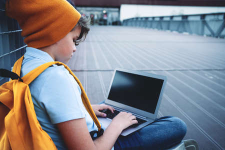 Child caucasian teen boy in yellow beany hat and backpack sitting outdoors using laptop. Remote or...