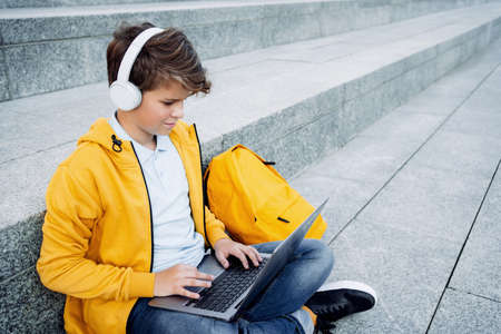 Child caucasian teen boy in yellow hoodie with headphones sitting outdoors using laptop. Remote or distant learning on the go. Blogging surfing. Homeschooling. Lockdown. Video call. Copy space.の写真素材