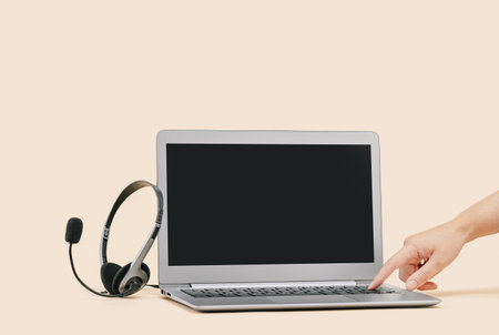 Front view of slim laptop with headset on beige. Female hand turning on. Distant learning. working from home, online courses or support. Audio podcast headphones . Helpdesk or call center bannerの写真素材