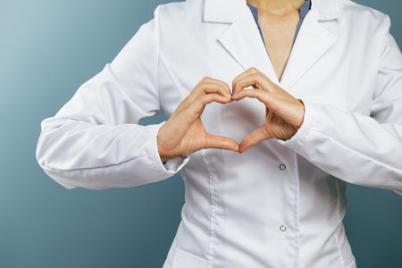 Female doctor in white uniform forms a heart shape with her hands. Minimal on blue background. Banner copy space. Heart, cardiology and medical care support and assistance female health gynecologyの写真素材