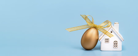 Happy Easter banner. One golden egg with a ribbon bow and white ceramic house on blue. Copy spaceの写真素材