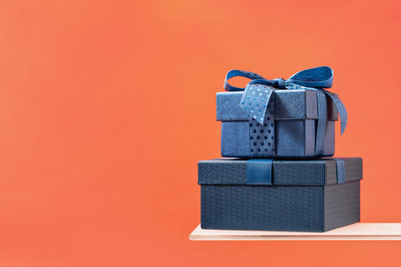 Two Luxury gift boxes with a blue bow on coral background. Side view on shelf. Fathers day or Valentines day gift for him. Corporate gift concept or birthday party. Festive sale copy space bannerの写真素材