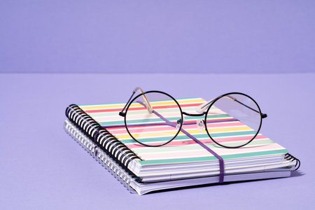 Eyeglasses on stripy notepad notebook on lilac purple desk. Back to School stationery. Spectacles or...