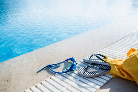 Blue Goggles flip flops and yellow beach towel on the side of swimming pool or seaside. Water and sand. Sunlit. Summer leisure activity and sports concept. Copy spaceの写真素材
