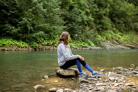 Young sports woman sitting on stone near the river and looking in the mountains, sports consept