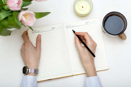 Woman hands holding planner on white, office desk with notebook, coffee, tulips, flat lay top view....