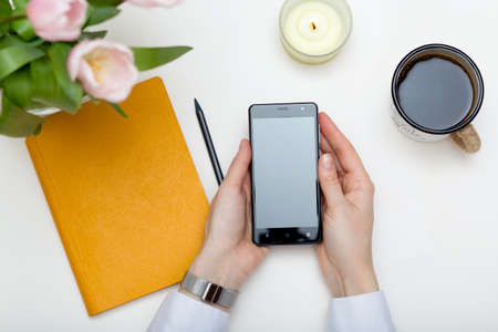 Women's hands holding up-to-date smartphone, white background. Coffee break top view. Women's business concept. Mockup for your design.の写真素材