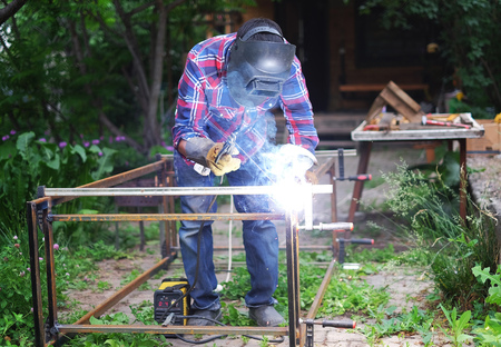 Man in a protective mask welding metal construction outside the house on a summer day
