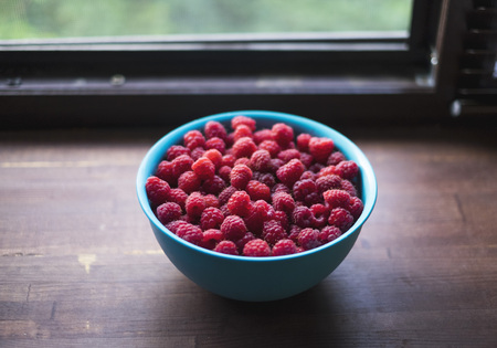 Blue bowl with fresh raspberries near the windowの写真素材