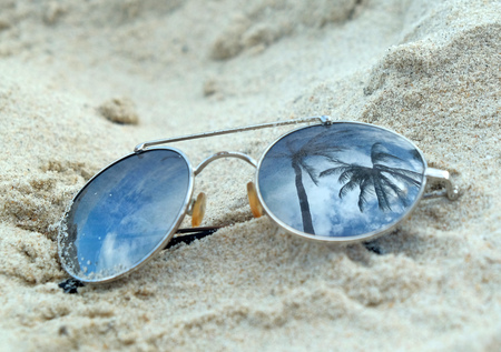 Mirrored sunglasses close up on the beach sand with palm trees reflection.