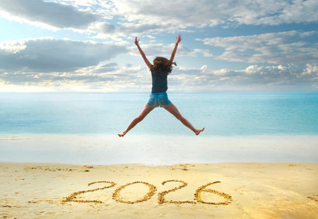2026 written on sand, happy girl with hands up jumping on the beach.の写真素材