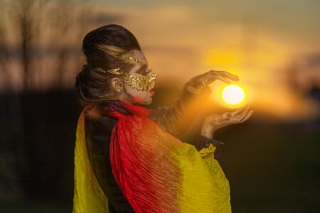 Young woman in a golden mask spikes on her face dancing with fans and shawls and posing at sunset