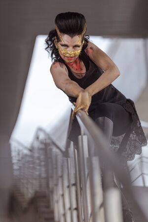 Young woman in a golden mask spikes on her face in a black dress and boots posing near modern buildings urban cityの写真素材