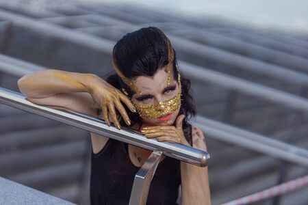 Young woman in a golden mask spikes on her face in a black dress and boots posing near modern buildings urban cityの写真素材