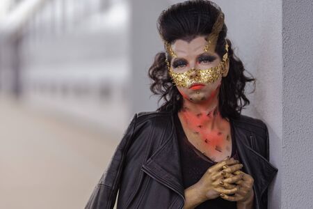 Young woman in a golden mask spikes on her face in a black dress and boots posing near modern...