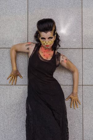 Young woman in a golden mask spikes on her face in a black dress and boots posing near modern...