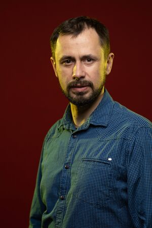 A brunette man with a beard in a blue shirt poses on a dark red background in the studio