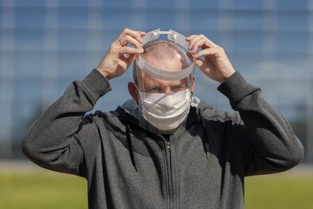 adult bald man wearing a white protective mask puts on a protective transparent visor on his face on...
