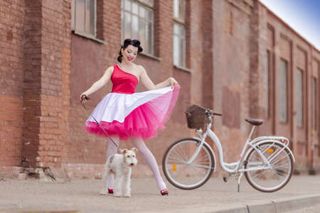 A girl in a dress and hairstyle in the style of the 40-50s on the city street with a dog breed Fox Terrier on a leash on a sunny day. Retro style photo.の写真素材