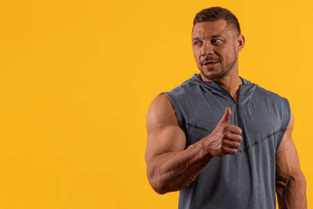Emotional young male athlete bodybuilder in a gray T-shirt posing on a yellow background