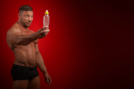 Young male athlete bodybuilder nude torso holding a bottle of drinking water in his hands and posing...