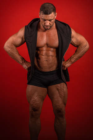 Young male athlete bodybuilder in a black sleeveless vest and shorts posing on a red background