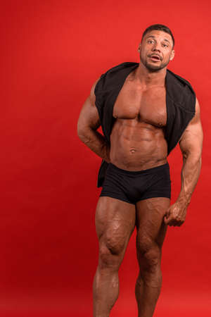 Young male athlete bodybuilder in a black sleeveless vest and shorts posing on a red backgroundの写真素材