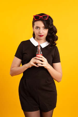 Pinup young brunette girl in a short black dress and red glasses holds a bottle with a drink in her hands and poses on a yellow backgroundの写真素材