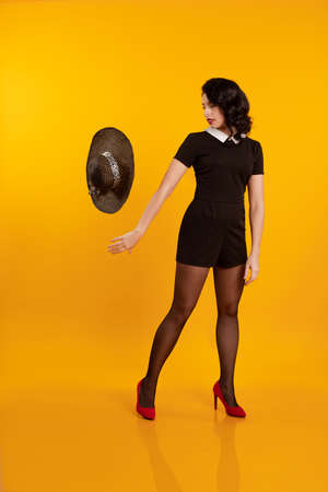 Young brunette girl in a short black dress holds a black hat with large brim and poses on a yellow...