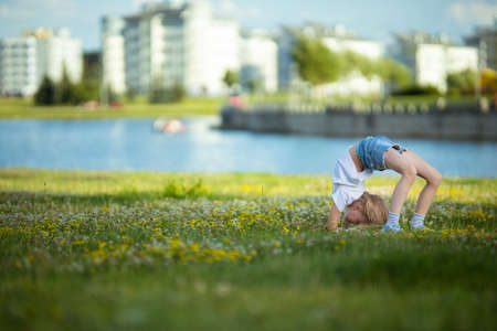 Blonde girl dancing, jumping, doing acrobatics and posing, in a city park on green grass near a lake on a sunny dayの写真素材