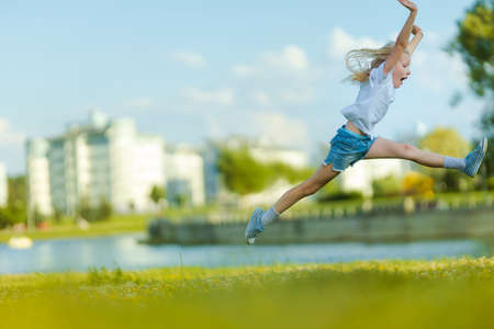 Blonde girl dancing, jumping, doing acrobatics and posing, in a city park on green grass near a lake on a sunny dayの写真素材