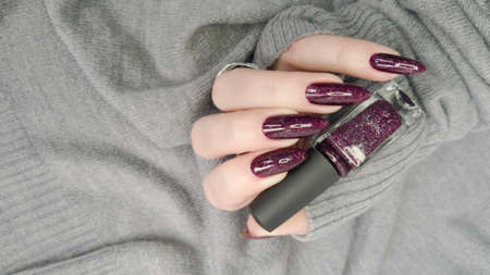 Woman's hands with long nails and a dark red burgundy manicure holds a bottle of nail polish