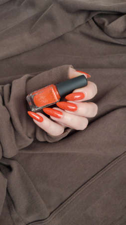 Woman hand with long nails and orange ginger manicure holds a bottle of nail polish