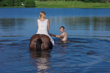 Young blonde girl in a white long dress swims in the lake with a brown horse on a sunny dayの写真素材