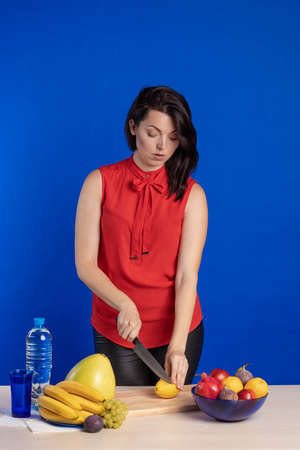 Emotional actress woman in a red blouse posing with a knife and lemon fruit in her hands on a blue...