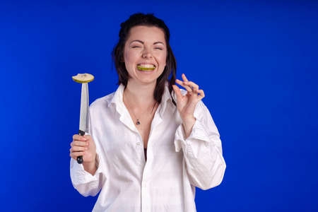 Emotional actress woman in a white shirt posing with fruits and a knife in her hands and on the table on a blue backgroundの写真素材