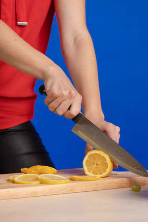 Woman in red blouse slices lemon fruit on a wooden board on a table with a large cook knife on a blue backgroundの写真素材