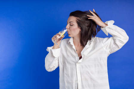 Emotional young woman in a white shirt poses with a glass of clean drinking water and a slice of lemon in her hands on a blue backgroundの写真素材