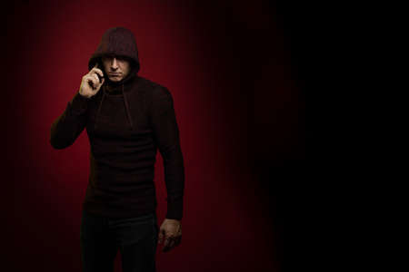A man in a jumper with a hood and with a phone in his hands poses against a dark red backgroundの写真素材