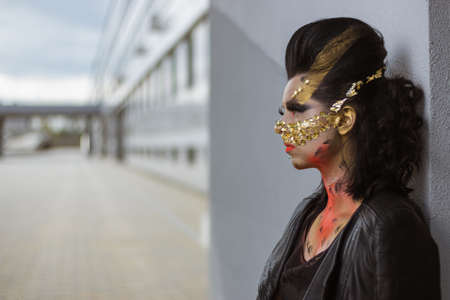 Actress woman in a golden mask spikes on her face in a black dress posing near modern buildings urban cityの写真素材