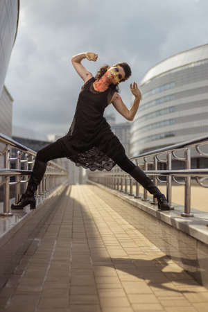 Actress woman in a golden mask spikes on her face in a black dress posing near modern buildings urban cityの写真素材