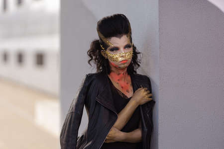 Actress woman in a golden mask spikes on her face in a black dress posing near modern buildings...