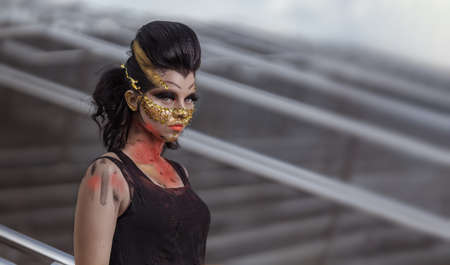 Actress woman in a golden mask spikes on her face in a black dress posing near modern buildings urban cityの写真素材