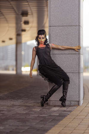 Actress woman in a golden mask spikes on her face in a black dress posing near modern buildings...
