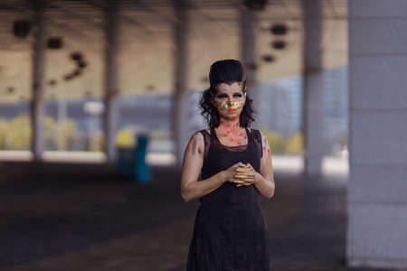 Actress woman in a golden mask spikes on her face in a black dress posing near modern buildings...