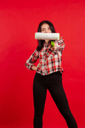 Girl master builder painter in a plaid shirt with a roller for painting poses on a red background