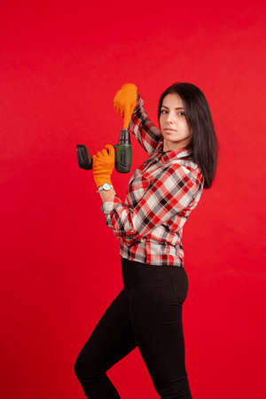 Builder girl in a plaid shirt and orange gloves holds an electric drill and poses on a red backgroundの写真素材