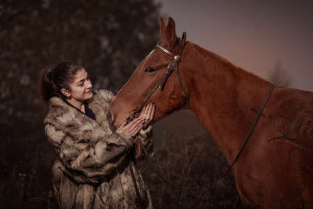 A young brunette woman in a gray coat poses with a brown horse. Cold autumn in the forest on the...